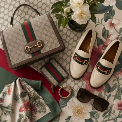 GUCCI — ICONIC ITALIAN LUXURY, DELIVERED WORLDWIDE featuring a Gucci monogram handbag, matching loafers, silk scarf, sunglasses, and jewelry arranged in an elegant floral setting, showcasing Italian luxury style.
