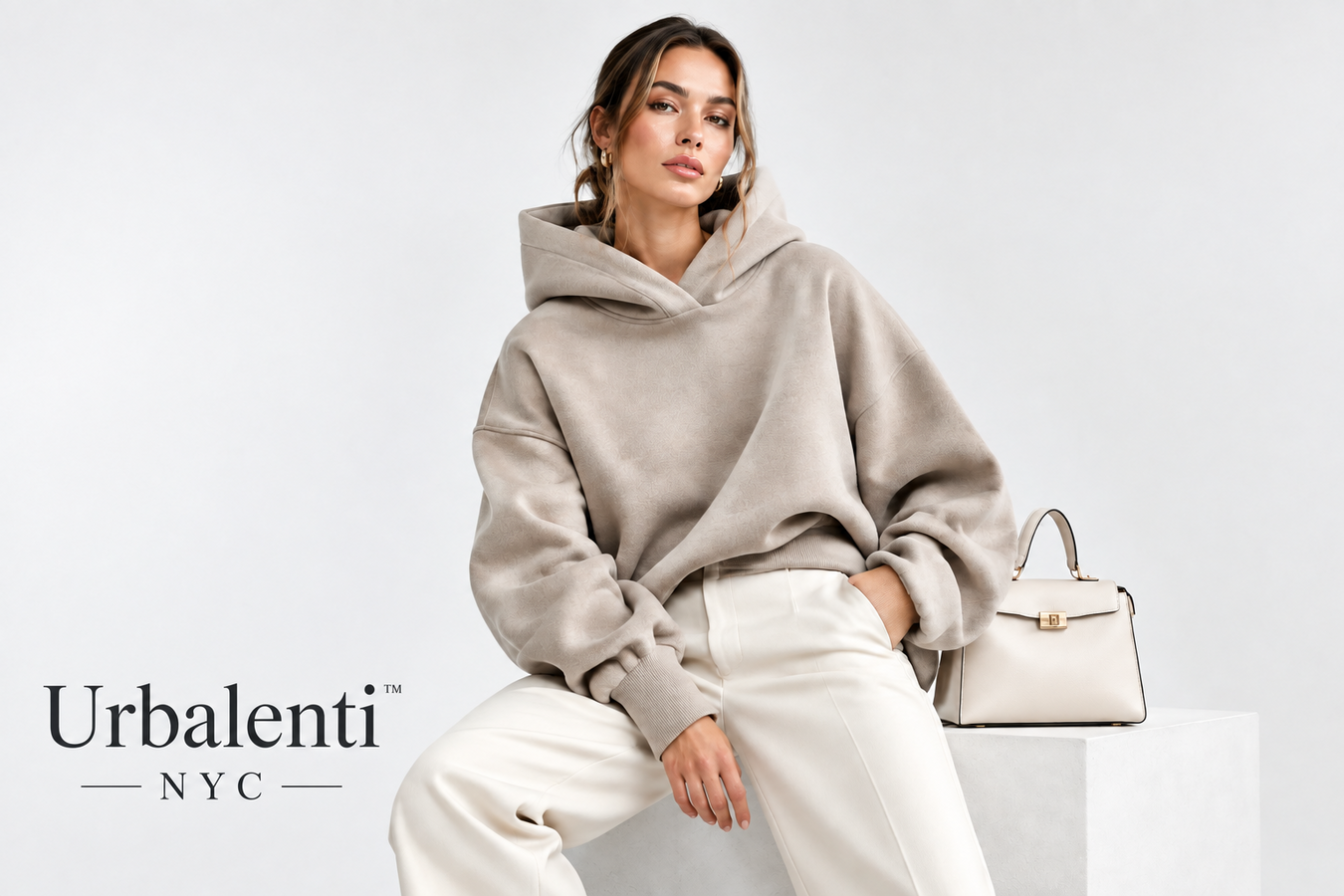 Woman wearing a taupe oversized hoodie with white pants and a handbag, showcasing Designer Hoodies for Women. Shop Designer Hoodies for Women Now at Urbalenti NYC.