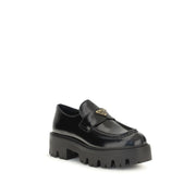 Prada Black Brushed Leather Slip-On Loafers for Women with Triangle Logo, angled front view highlighting the enameled Prada triangle logo, leather vamp strap, moc-toe stitching, and lugged rubber sole. Prada Loafers for Women from Urbalenti NYC, style 13879, SKU 79661334579, Black, Discount_30-39, available in EU36.5/US6.5, EU36/US6, EU37/US7, EU38.5/US8.5, EU39/US9, and EU40/US10; in-stock, Loafers - Shoes, New with tags, Prada, Shoes, Slip-On Loafers - Loafers - Shoes, Women.