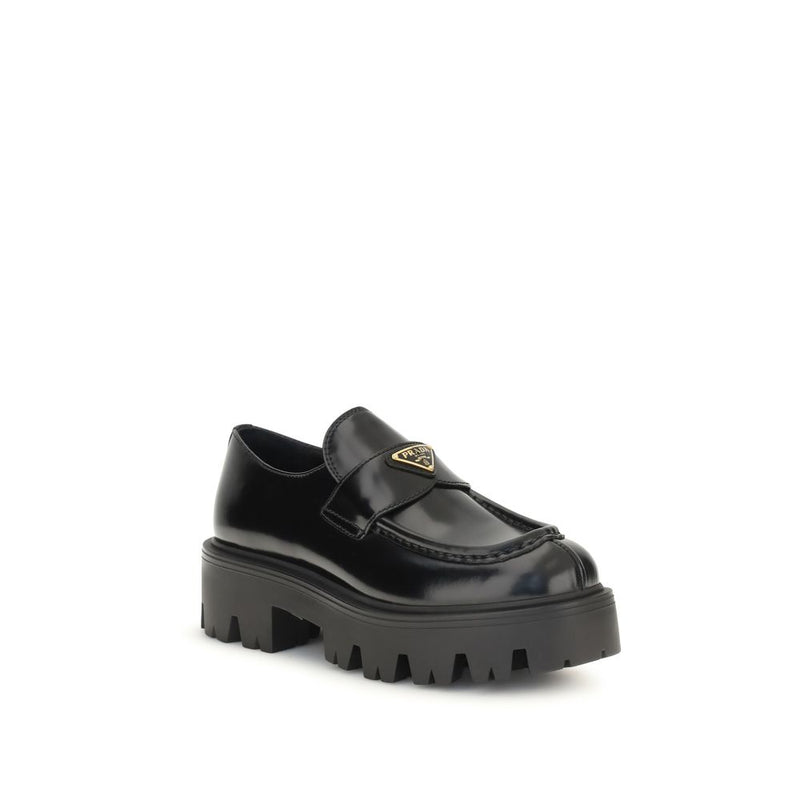 Prada Black Brushed Leather Slip-On Loafers for Women with Triangle Logo, angled front view highlighting the enameled Prada triangle logo, leather vamp strap, moc-toe stitching, and lugged rubber sole. Prada Loafers for Women from Urbalenti NYC, style 13879, SKU 79661334579, Black, Discount_30-39, available in EU36.5/US6.5, EU36/US6, EU37/US7, EU38.5/US8.5, EU39/US9, and EU40/US10; in-stock, Loafers - Shoes, New with tags, Prada, Shoes, Slip-On Loafers - Loafers - Shoes, Women.