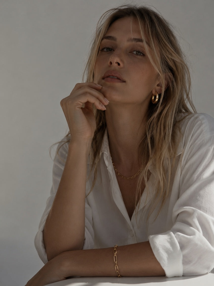 A woman with blonde hair wearing a white button-down shirt, gold hoop earrings, and a delicate gold chain necklace and bracelet, resting her chin on her hand against a neutral light gray background.